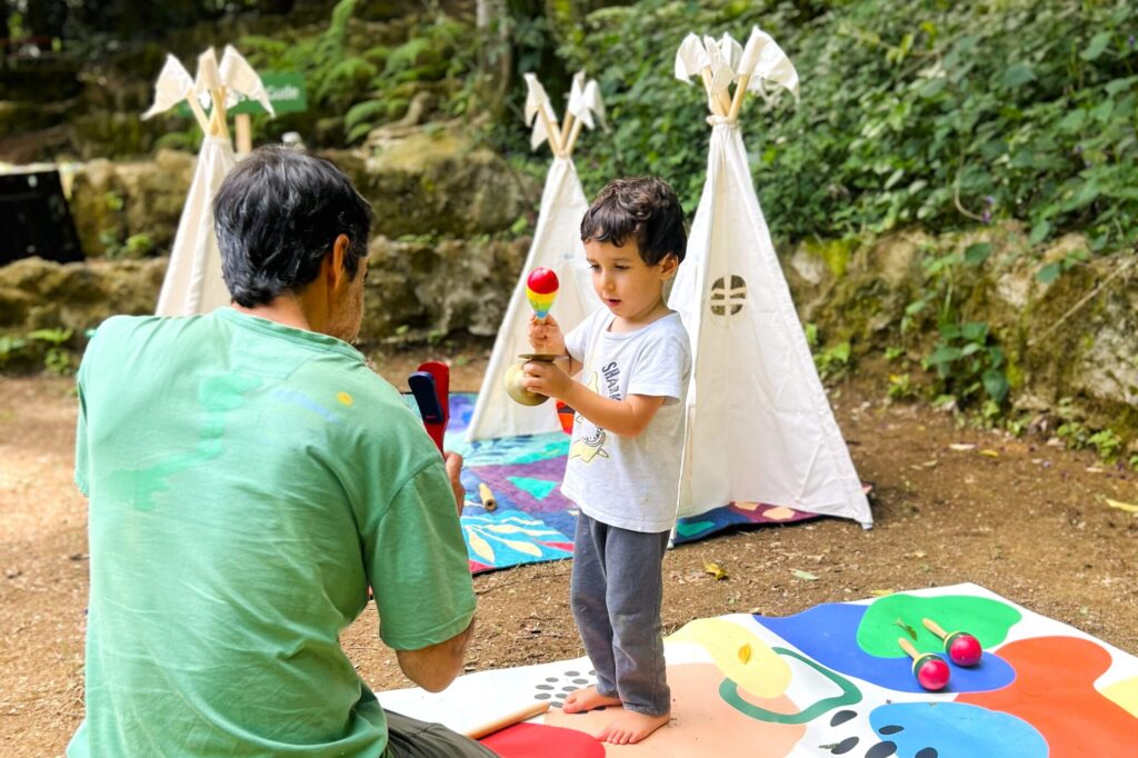 Criança brincando com instrumentos musicais em atividade ao ar livre, acompanhada por um adulto em um espaço cercado pela natureza durante uma festa infantil.